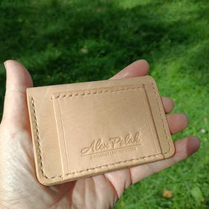 May include: A light tan leather card holder with visible stitching along the edges and a rectangular pocket on the front. The leather is embossed with the text Alex Polak HANDMADE LEATHER GOODS in a centered, stylized font on the front pocket.