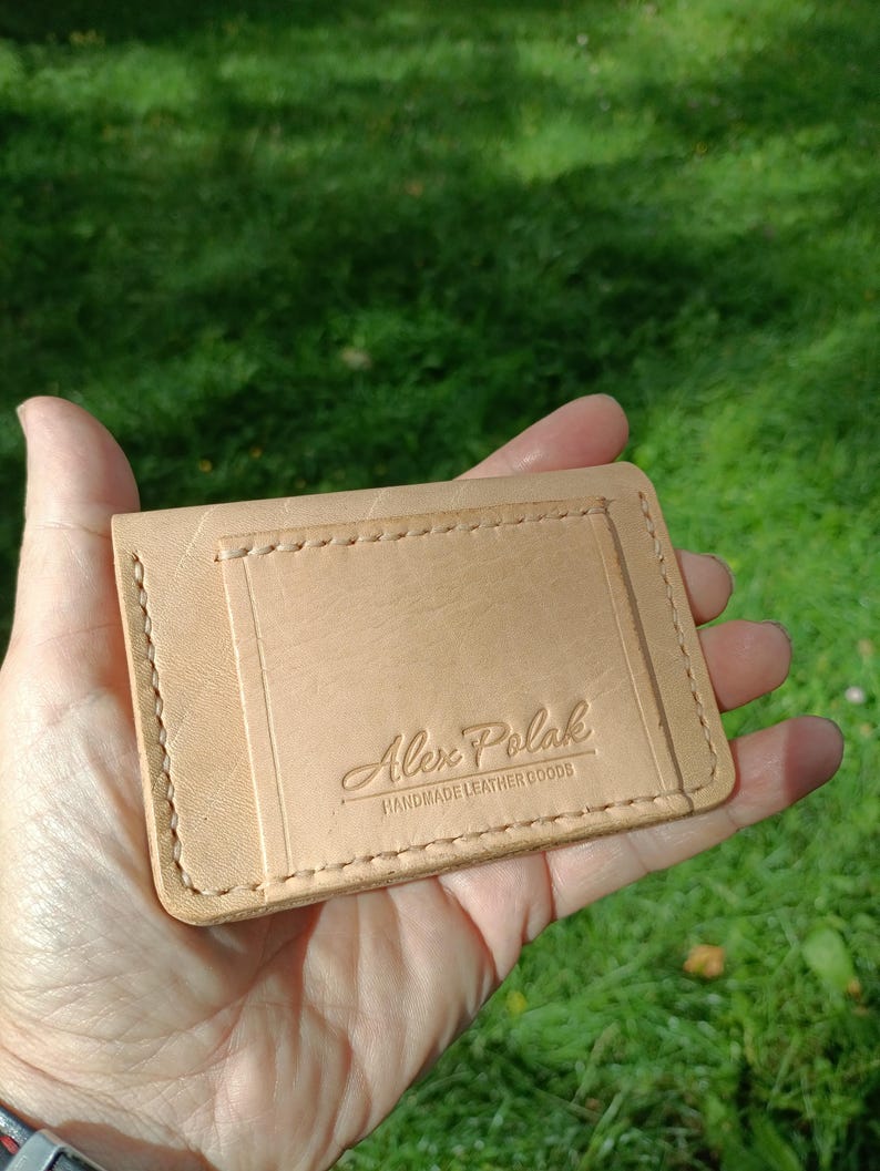 May include: A light tan leather card holder with visible stitching along the edges and a rectangular pocket on the front. The leather is embossed with the text Alex Polak HANDMADE LEATHER GOODS in a centered, stylized font on the front pocket.