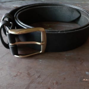 May include: A black belt featuring a smooth, matte finish and a rectangular, metallic buckle with a brushed silver tone. The belt includes two matching black leather loops near the buckle, which is secured by a single prong mechanism.
