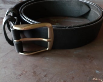 May include: A black belt featuring a smooth, matte finish and a rectangular, metallic buckle with a brushed silver tone. The belt includes two matching black leather loops near the buckle, which is secured by a single prong mechanism.