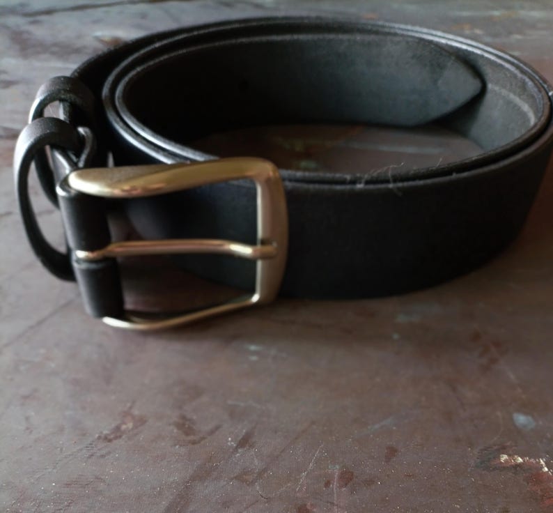 Puede incluir: Un cintur&oacute;n negro con acabado liso y mate, que presenta una hebilla met&aacute;lica rectangular en tono plateado cepillado. El cintur&oacute;n incluye dos trabillas de cuero negro a juego cerca de la hebilla, asegurada mediante un mecanismo de un solo perno.