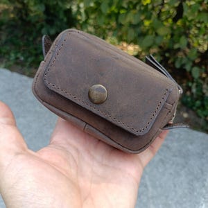 Peut inclure: Une petite pochette rectangulaire en cuir marron dot&eacute;e d'un rabat ferm&eacute; par un bouton-pression en m&eacute;tal couleur laiton vieilli. La pochette pr&eacute;sente des bords cousus visibles et une surface mate textur&eacute;e, con&ccedil;ue pour le rangement d'objets.
