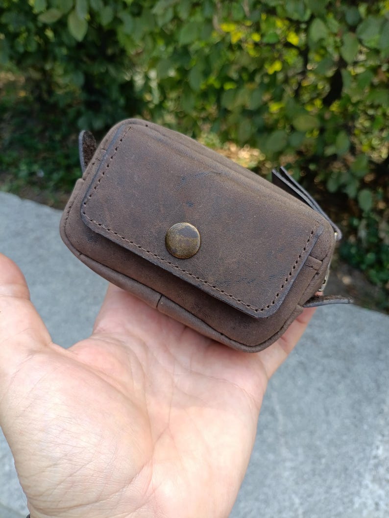 Peut inclure: Une petite pochette rectangulaire en cuir marron dot&eacute;e d'un rabat ferm&eacute; par un bouton-pression en m&eacute;tal couleur laiton vieilli. La pochette pr&eacute;sente des bords cousus visibles et une surface mate textur&eacute;e, con&ccedil;ue pour le rangement d'objets.