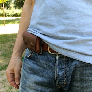 Puede incluir: Un cintur&oacute;n de cuero marr&oacute;n con una funda rectangular sujeta en el lateral. La funda se asegura con dos broches met&aacute;licos de color lat&oacute;n. El cintur&oacute;n tiene una hebilla de metal plateado y se lleva sobre unos vaqueros azules y una camiseta azul claro.