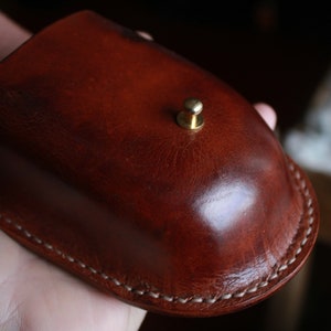 May include: A brown leather pouch with a brass button closure. The pouch has a white stitching detail around the edges.