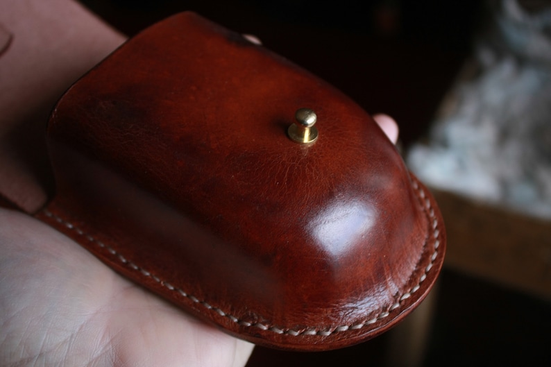 May include: A brown leather pouch with a brass button closure. The pouch has a white stitching detail around the edges.
