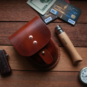 May include: A brown leather pouch with a snap closure, a pocket knife with a wooden handle, a pocket watch with a silver chain, and a lighter with a brown leather case.
