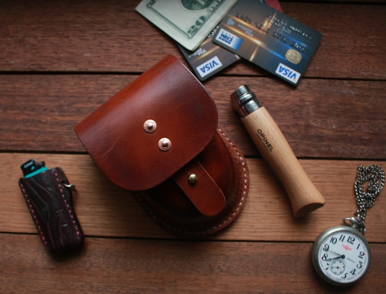May include: A brown leather pouch with a snap closure, a pocket knife with a wooden handle, a pocket watch with a silver chain, and a lighter with a brown leather case.