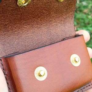 May include: A brown leather pouch featuring a fold-over flap with two gold-toned metal snap buttons. The interior of the flap shows the textured underside of the leather, while the front exterior is smooth. Two matching gold snap studs are visible.