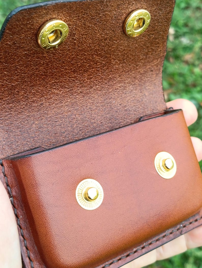 May include: A brown leather pouch featuring a fold-over flap with two gold-toned metal snap buttons. The interior of the flap shows the textured underside of the leather, while the front exterior is smooth. Two matching gold snap studs are visible.