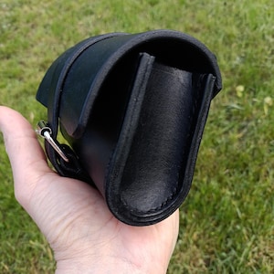 May include: A black leather bag with a curved design, featuring a strap and a silver-colored buckle. The bag is held against a backdrop of green grass.