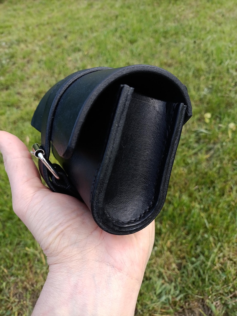 May include: A black leather bag with a curved design, featuring a strap and a silver-colored buckle. The bag is held against a backdrop of green grass.
