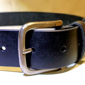 Op de afbeelding: Close-up van een zwarte leren riem met een zilverkleurige gesp. De gesp heeft een rechthoekig frame en een goudkleurige staaf. De riem heeft een enkele lus en verstelgaten. De riem is ontworpen voor dagelijks gebruik.