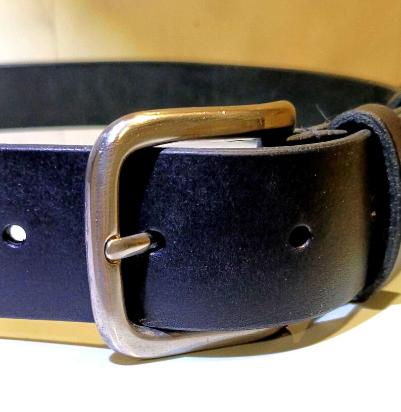 Op de afbeelding: Close-up van een zwarte leren riem met een zilverkleurige gesp. De gesp heeft een rechthoekig frame en een goudkleurige staaf. De riem heeft een enkele lus en verstelgaten. De riem is ontworpen voor dagelijks gebruik.