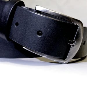 May include: A close-up shot of a black leather belt with a silver buckle. The belt features a classic design with a rectangular buckle and a single loop. The leather looks smooth and the buckle is made of metal.