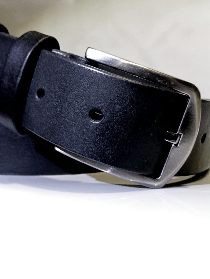 May include: A close-up shot of a black leather belt with a silver buckle. The belt features a classic design with a rectangular buckle and a single loop. The leather looks smooth and the buckle is made of metal.
