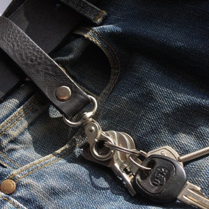 May include: A black leather belt with a silver key chain attached to it. The belt is fastened to a pair of blue denim jeans.