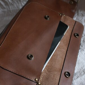 May include: A brown leather satchel bag with a flap closure and a pocket on the front. The bag has a zippered pocket on the inside and a strap for carrying.