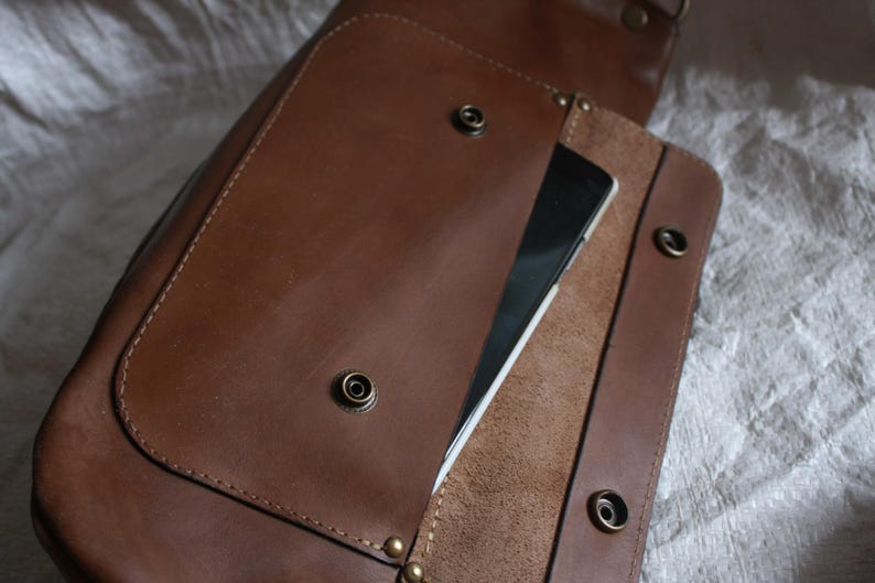 May include: A brown leather satchel bag with a flap closure and a pocket on the front. The bag has a zippered pocket on the inside and a strap for carrying.