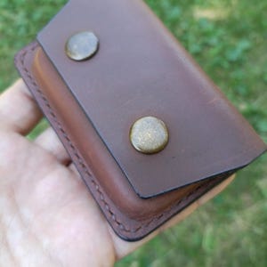 Puede incluir: Una funda rectangular de cuero marr&oacute;n con cierre de solapa asegurado por dos broches met&aacute;licos redondos en tono lat&oacute;n. Los bordes de la funda tienen costuras oscuras visibles y el cuero presenta una textura de superficie lisa y mate.