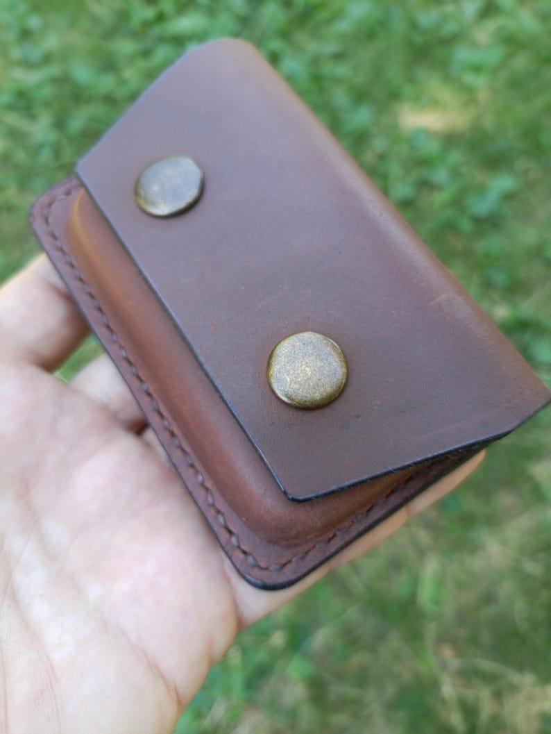 Puede incluir: Una funda rectangular de cuero marr&oacute;n con cierre de solapa asegurado por dos broches met&aacute;licos redondos en tono lat&oacute;n. Los bordes de la funda tienen costuras oscuras visibles y el cuero presenta una textura de superficie lisa y mate.