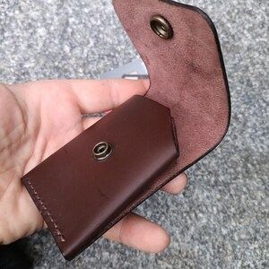 May include: A dark brown leather card holder featuring a flap closure with a brass-colored metal snap button. The interior of the leather is visible, showing a natural suede texture, and the exterior is smooth with visible stitching along the side.
