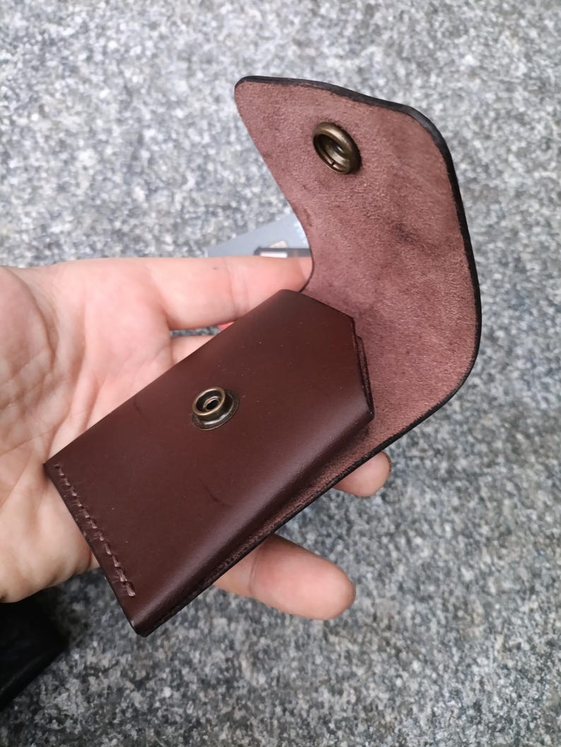 May include: A dark brown leather card holder featuring a flap closure with a brass-colored metal snap button. The interior of the leather is visible, showing a natural suede texture, and the exterior is smooth with visible stitching along the side.