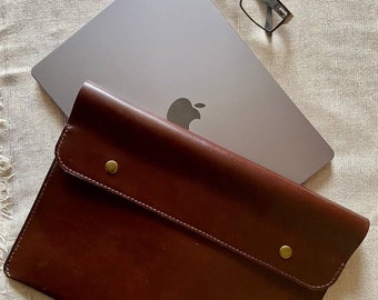 May include: A brown leather laptop sleeve with a flap closure and a silver laptop with an Apple logo inside. The sleeve has two brass rivets on the flap. 