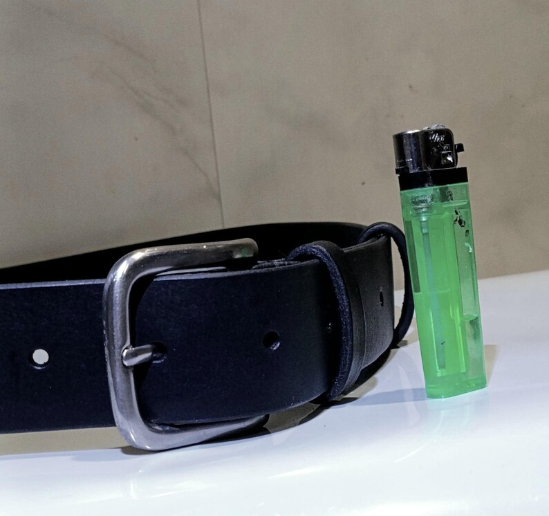 Op de afbeelding: Een zwarte leren riem met een zilverkleurige gesp en een groene aansteker. De riem is gemaakt van glad leer en heeft een klassiek ontwerp. De aansteker is transparant met een zwarte bovenkant.