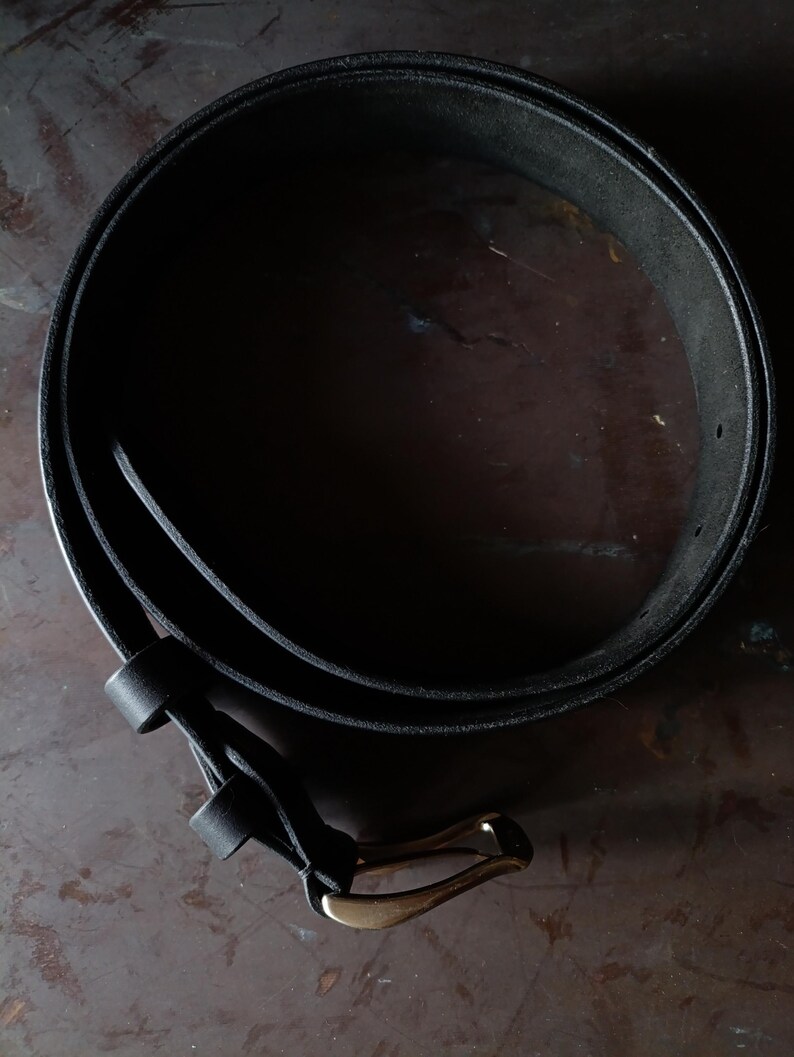 Puede incluir: Un cintur&oacute;n de cuero negro enrollado en forma circular, con un acabado mate liso y una hebilla met&aacute;lica en tono lat&oacute;n. El cintur&oacute;n incluye una trabilla de cuero cerca de la hebilla y orificios de ajuste visibles a lo largo de la tira.