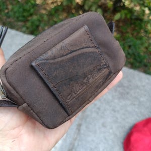 Peut inclure: Une petite pochette rectangulaire marron dot&eacute;e d'un empi&egrave;cement en cuir textur&eacute; sur le devant avec un logo en relief. La pochette est fabriqu&eacute;e dans un tissu marron fonc&eacute; mat et comprend une fermeture &eacute;clair en m&eacute;tal sur le bord sup&eacute;rieur.