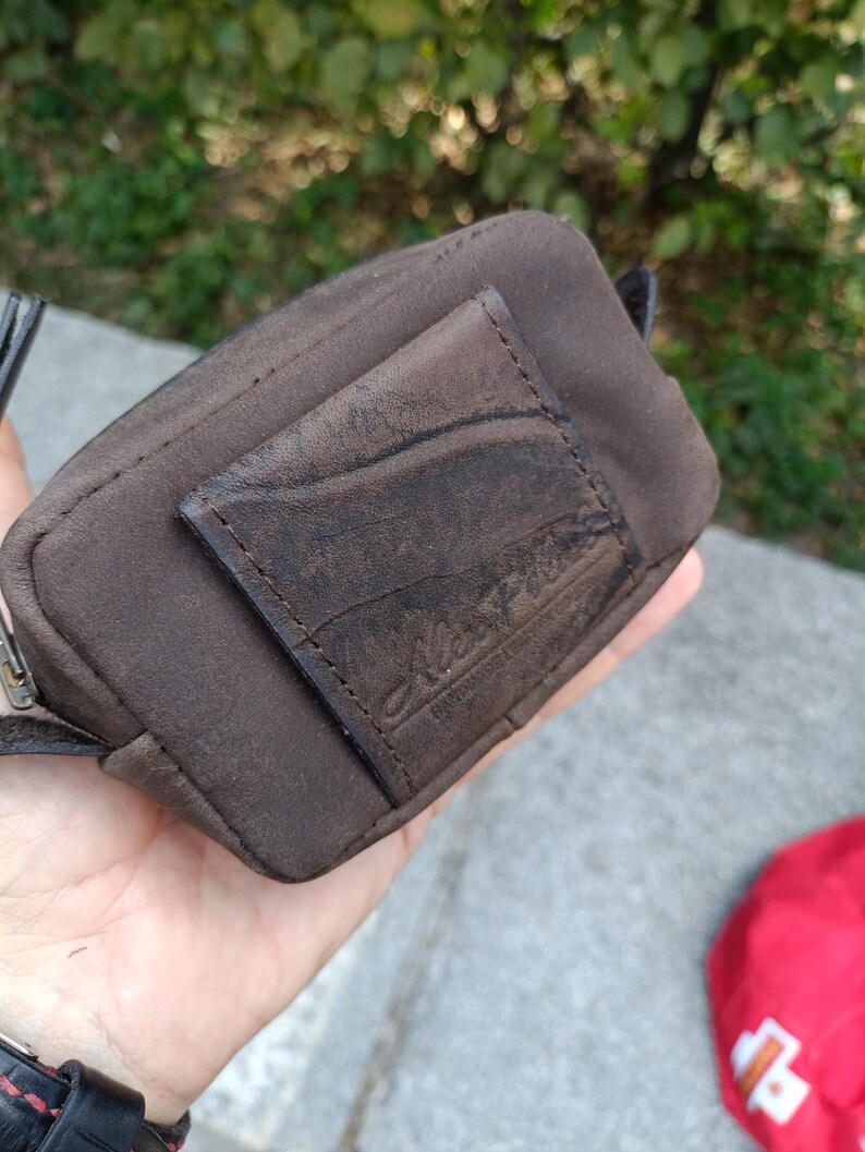 Peut inclure: Une petite pochette rectangulaire marron dot&eacute;e d'un empi&egrave;cement en cuir textur&eacute; sur le devant avec un logo en relief. La pochette est fabriqu&eacute;e dans un tissu marron fonc&eacute; mat et comprend une fermeture &eacute;clair en m&eacute;tal sur le bord sup&eacute;rieur.
