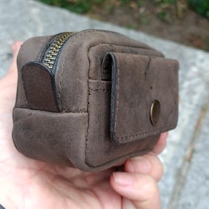 Peut inclure: Une petite pochette rectangulaire en cuir marron avec une finition mate et textur&eacute;e. La pochette comprend une fermeture &eacute;clair sur le dessus avec une tirette en m&eacute;tal fonc&eacute; et une poche ext&eacute;rieure avant s&eacute;curis&eacute;e par un rabat &agrave; bouton-pression.