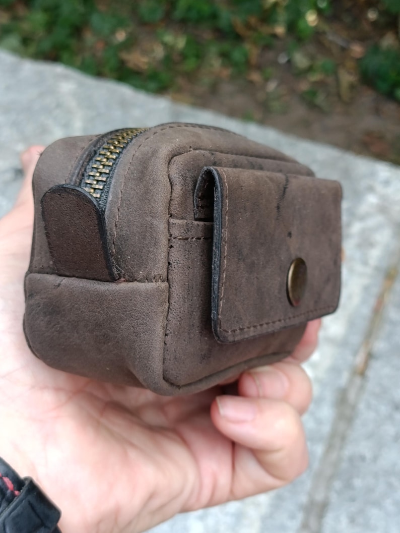 Peut inclure: Une petite pochette rectangulaire en cuir marron avec une finition mate et textur&eacute;e. La pochette comprend une fermeture &eacute;clair sur le dessus avec une tirette en m&eacute;tal fonc&eacute; et une poche ext&eacute;rieure avant s&eacute;curis&eacute;e par un rabat &agrave; bouton-pression.