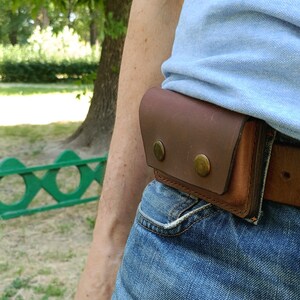 Puede incluir: Una funda de cintur&oacute;n de cuero marr&oacute;n con una solapa rectangular asegurada por dos botones de presi&oacute;n met&aacute;licos de color lat&oacute;n. La funda est&aacute; sujeta a un cintur&oacute;n de cuero marr&oacute;n que se lleva sobre unos vaqueros azules y una camisa azul claro.