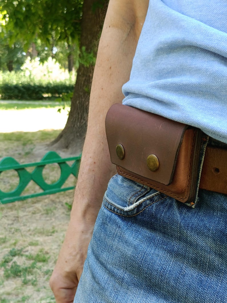 Puede incluir: Una funda de cintur&oacute;n de cuero marr&oacute;n con una solapa rectangular asegurada por dos botones de presi&oacute;n met&aacute;licos de color lat&oacute;n. La funda est&aacute; sujeta a un cintur&oacute;n de cuero marr&oacute;n que se lleva sobre unos vaqueros azules y una camisa azul claro.
