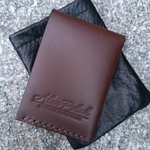 May include: A dark brown leather card holder with visible stitching along the bottom edge and the embossed text Alex Polak HANDMADE LEATHER GOODS, resting on a black textured leather pouch with a drawstring closure.