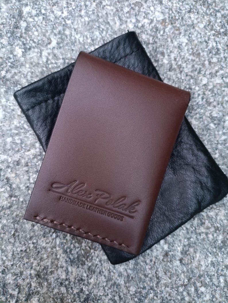 May include: A dark brown leather card holder with visible stitching along the bottom edge and the embossed text Alex Polak HANDMADE LEATHER GOODS, resting on a black textured leather pouch with a drawstring closure.
