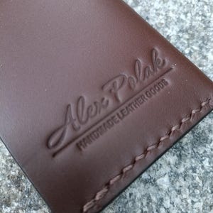 May include: A brown leather wallet featuring a debossed logo that reads Alex Polak HANDMADE LEATHER GOODS. The wallet is constructed with visible brown stitching along the edge, showcasing a smooth surface texture and a clean, rectangular design.