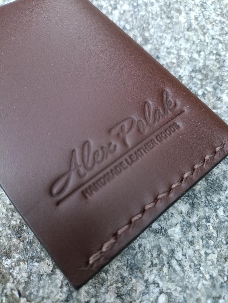 May include: A brown leather wallet featuring a debossed logo that reads Alex Polak HANDMADE LEATHER GOODS. The wallet is constructed with visible brown stitching along the edge, showcasing a smooth surface texture and a clean, rectangular design.