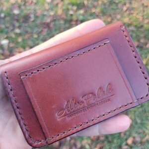 May include: A brown leather card holder featuring visible saddle stitching along the edges and a rectangular pocket on the front. The leather is embossed with the text Alex Polak HANDMADE LEATHER GOODS in a script and block font combination.
