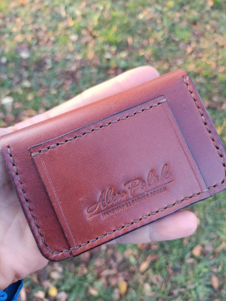 May include: A brown leather card holder featuring visible saddle stitching along the edges and a rectangular pocket on the front. The leather is embossed with the text Alex Polak HANDMADE LEATHER GOODS in a script and block font combination.
