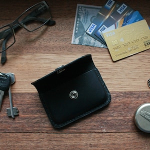 Puede incluir: Una cartera de cuero negro con cierre de presi&oacute;n. La cartera est&aacute; sobre una superficie de madera con unas gafas, llaves, un reloj de bolsillo y tarjetas de cr&eacute;dito.