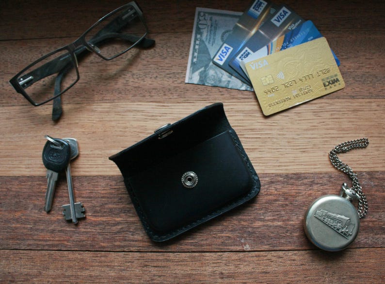 Puede incluir: Una cartera de cuero negro con cierre de presi&oacute;n. La cartera est&aacute; sobre una superficie de madera con unas gafas, llaves, un reloj de bolsillo y tarjetas de cr&eacute;dito.