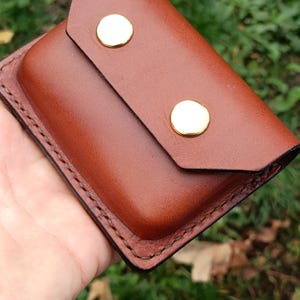 Puede incluir: Una cartera rectangular de cuero marr&oacute;n con un cierre de solapa asegurado por dos broches de metal dorado pulido. Los bordes de la cartera est&aacute;n terminados con costuras visibles en color marr&oacute;n oscuro, lo que le otorga una apariencia estructurada.