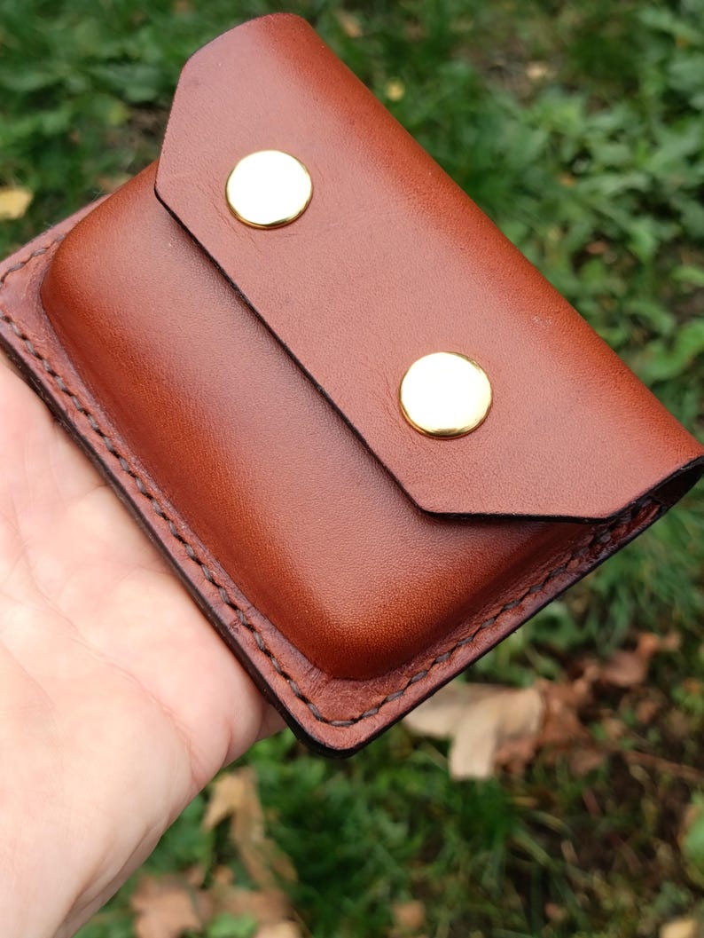Puede incluir: Una cartera rectangular de cuero marr&oacute;n con un cierre de solapa asegurado por dos broches de metal dorado pulido. Los bordes de la cartera est&aacute;n terminados con costuras visibles en color marr&oacute;n oscuro, lo que le otorga una apariencia estructurada.