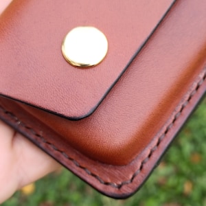 May include: A brown leather case featuring a smooth, rounded flap secured by a single polished gold-tone metal snap button. The edges of the case are finished with dark, visible stitching that follows the contour of the rectangular shape.
