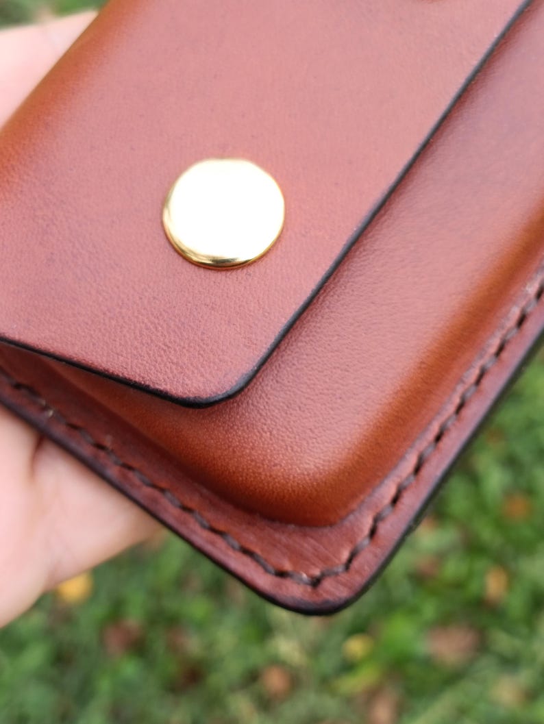 May include: A brown leather case featuring a smooth, rounded flap secured by a single polished gold-tone metal snap button. The edges of the case are finished with dark, visible stitching that follows the contour of the rectangular shape.