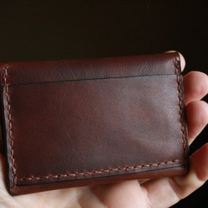 May include: A brown leather card holder with visible stitching. The wallet is rectangular and appears to be handmade. The leather has a rich, dark brown color and a smooth texture. The stitching is a lighter brown, providing contrast.