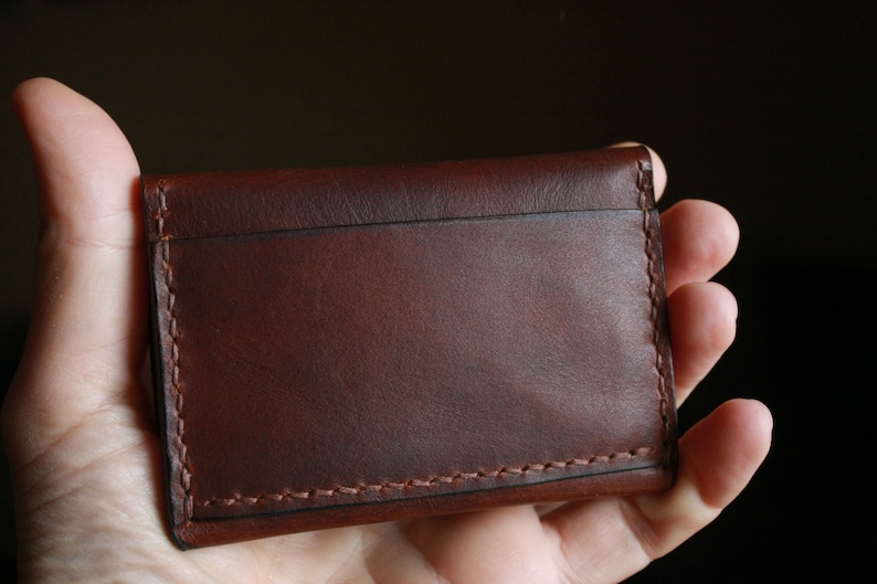 May include: A brown leather card holder with visible stitching. The wallet is rectangular and appears to be handmade. The leather has a rich, dark brown color and a smooth texture. The stitching is a lighter brown, providing contrast.